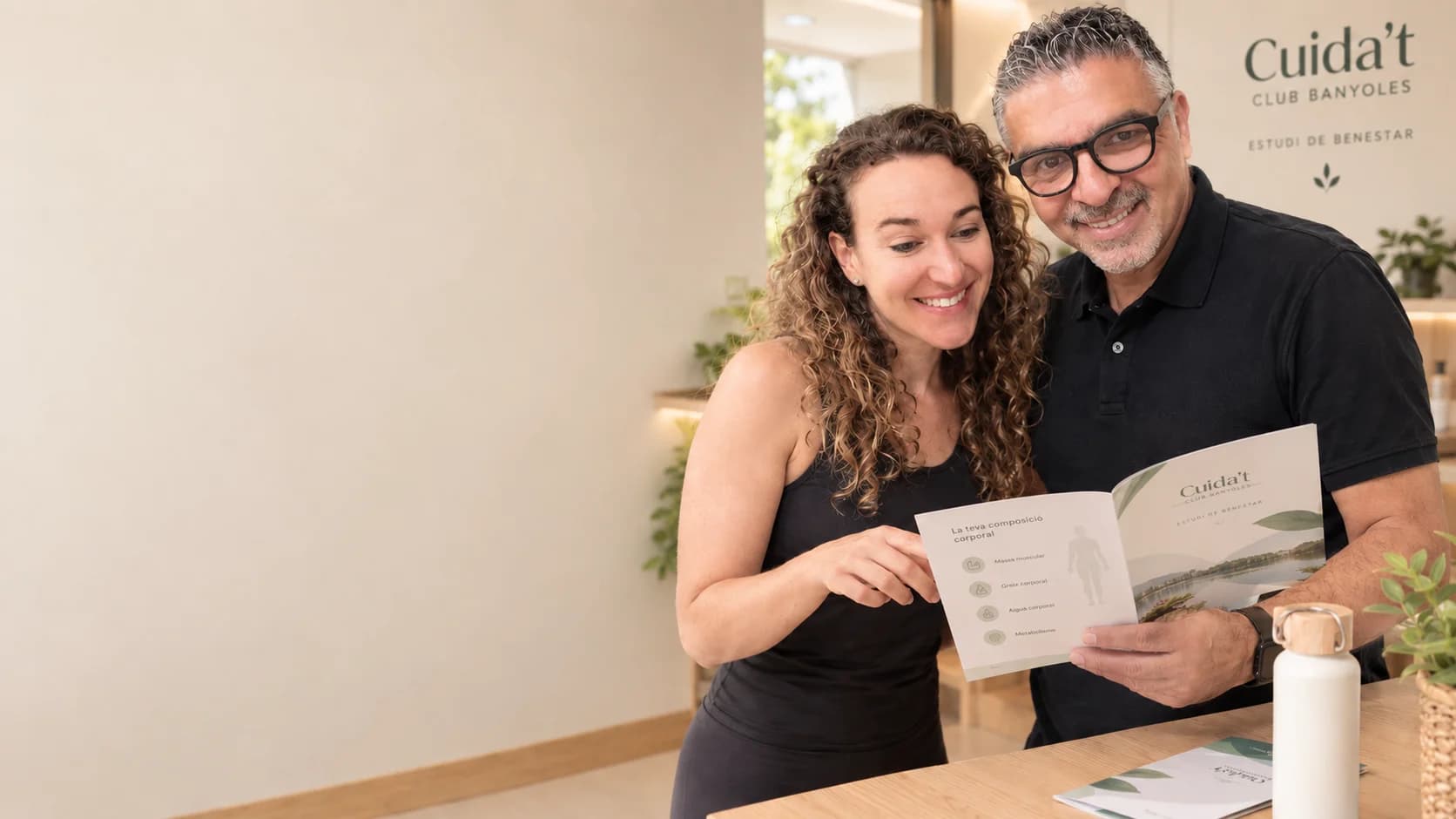 Dues persones somrient mentre revisen el fullet de l'Estudi de Benestar al local de Cuida't Club Banyoles